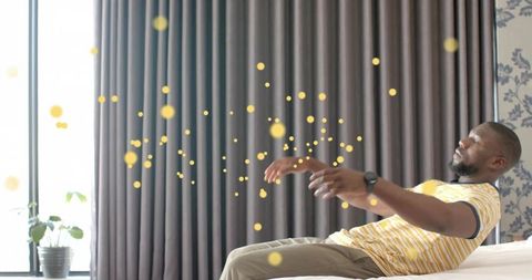 Relaxing man reclining on bed reaching toward glowing golden bokeh orbs in sunlit bedroom
