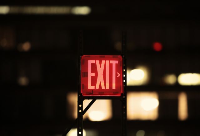 Glowing red exit sign useful for illustrating safety instructions, emergency exits, or indicating directions in buildings. The blurred city lights in the background create a sense of urban atmosphere and night-time setting, making it great for architectural or interior design visuals, advertisements for safety products, or urban nightlife illustrations.