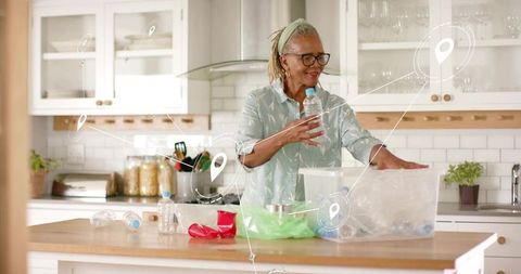 Senior woman sorting recyclables in modern kitchen placing plastic bottles into bin
