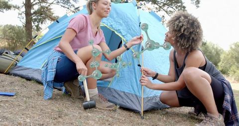 Two women assembling blue dome tent while camping outdoors, teamwork and hiking gear