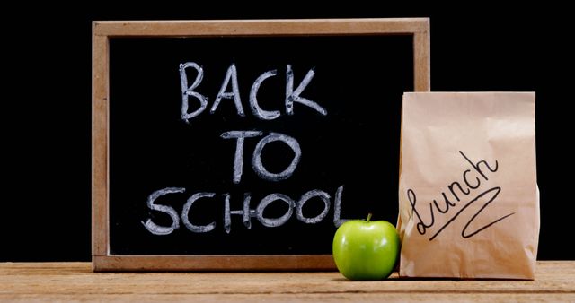 A chalkboard with the words Back to School written on it is displayed next to a green apple and a brown paper lunch bag, with copy space. It symbolizes the start of a new academic year and the preparation involved for students returning to their studies.