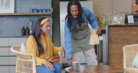 Friendly barista placing latte for customer in cozy wicker cafe with wood table and plants