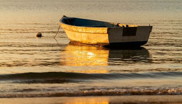 Weathered dinghy floating in golden sunrise reflection on calm shallow water with buoy