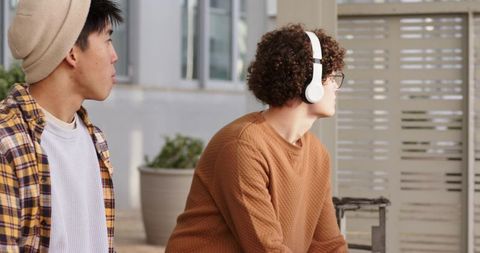 Two diverse young men sitting on bench listening to music with headphones outdoors