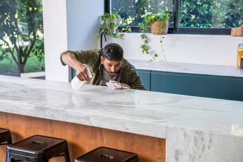 Mature indian man cleaning marble kitchen island with spray bottle and cloth, sunlit