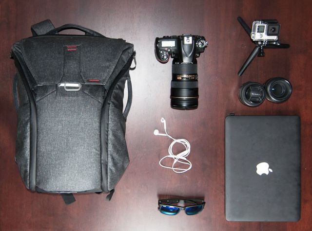 Staging travel essentials for photographers, this image features an organized display of a camera, a backpack, lenses, an Apple laptop, a GoPro, white earphones, and sunglasses. It highlights essential items for travel and photography, ideal for use in blogs or articles about travel packing, photography on the go, or organizing photography gear.