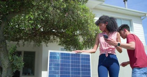 Multicultural couple installing solar panel in front yard, woman checking smartphone