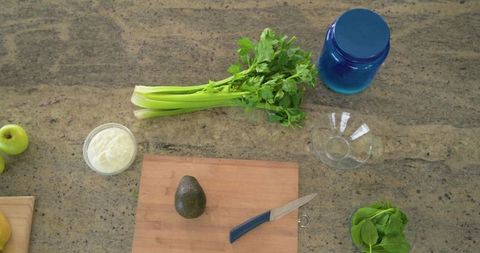 Overhead kitchen flatlay showing whole avocado on cutting board, celery and fresh greens