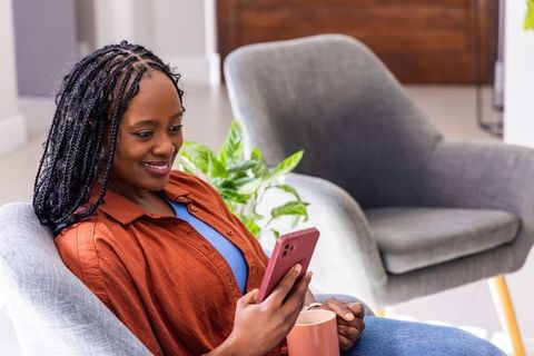 African woman relaxing in modern living room scrolling smartphone and enjoying coffee