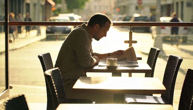 Man reading newspaper drinking coffee at sunlit sidewalk cafe
