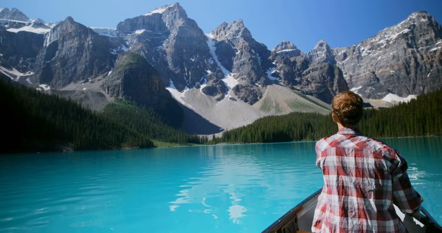 Individual wearing flannel shirt enjoys a tranquil canoeing experience on a pristine alpine lake with turquoise water. Majestic snow-capped mountains rise in the distance, while dense forests line the shores. Perfect for use in content related to outdoor activities, adventure, travel inspiration, and environmental conservation.