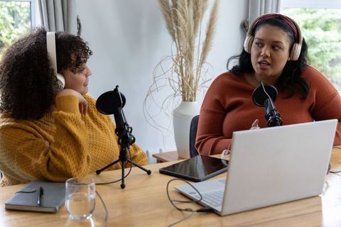 African american women hosting home podcast conversing with headphones, microphones, laptop