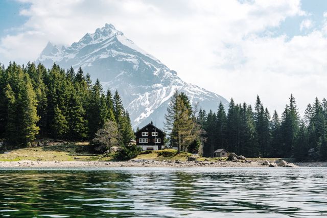 Cabin nestled in forest beside clear lake with snowy peaks in background, perfect for promoting travel, relaxation, wilderness retreats. Ideal for use in brochures, travel blogs, and advertisements emphasizing natural beauty and serene escapes.