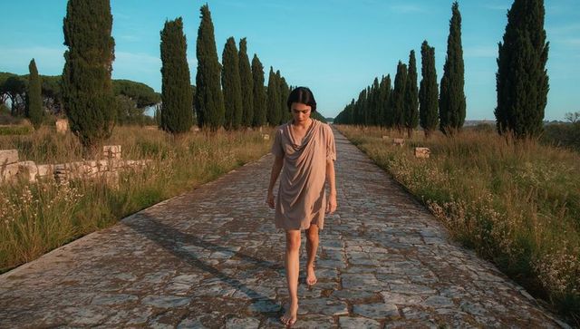 Barefoot woman walking along ancient stone avenue lined with tall cypress at golden hour