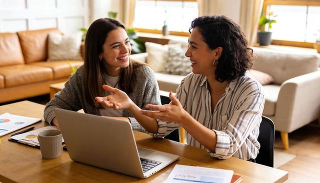 Two women collaborating at home desk with laptop and charts, smiling and brainstorming