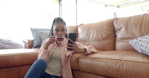 Relaxing woman sipping tea and scrolling smartphone by tan leather sofa in sunlit room
