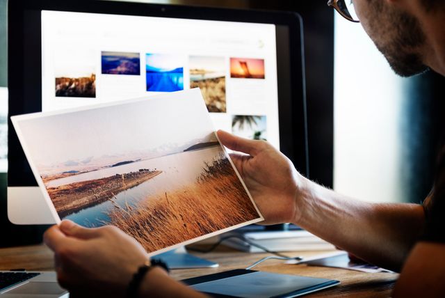 Photographer reviewing printed landscape photo at wooden desk. Background shows computer monitor displaying various images. Ideal for content related to photography work, digital editing, creative professionals, and office work environments.