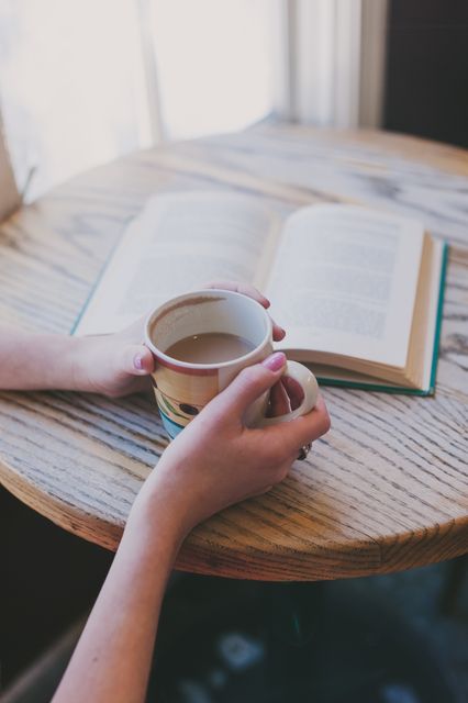 Hands holding coffee cup while an open book lies on wooden table. Chill vibes, perfect for literature, lifestyle, relaxation themes, and blog posts about hygge, self-care, quiet moments.