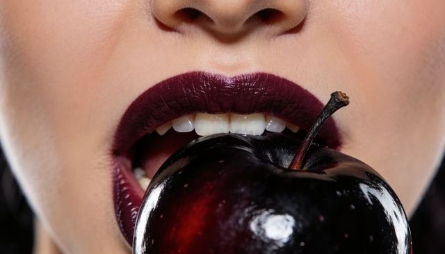 Close-up woman biting glossy dark-red apple wearing deep plum lipstick for beauty campaign