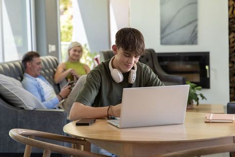 Teen working on laptop at home while family relaxing on sofa in cozy modern living room