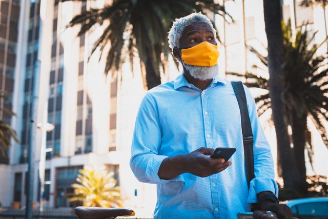 African american senior man wearing face mask using smartphone in the street. digital nomad on the go in the city during coronavirus covid 19 pandemic.