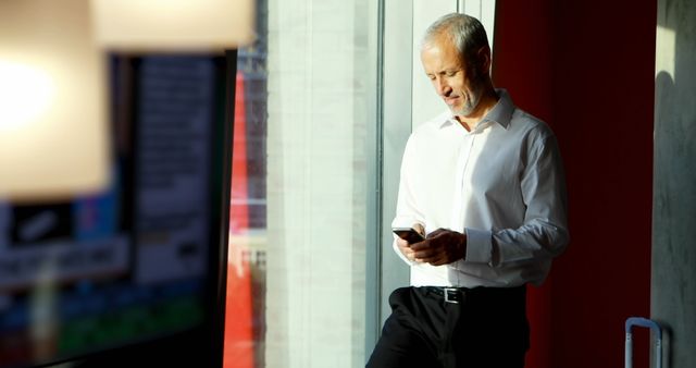 Ideal for business and professional contexts, this image shows a senior man reading his smartphone in a sunlit office. The man is wearing a white shirt, leaning against the wall, and looks concentrated and thoughtful. Perfect for depicting senior professionals in digital, tech-savvy settings, corporate culture, or retirement planning. Can be used in magazines, business reports, or websites related to business communication, technology, or modern work environments.