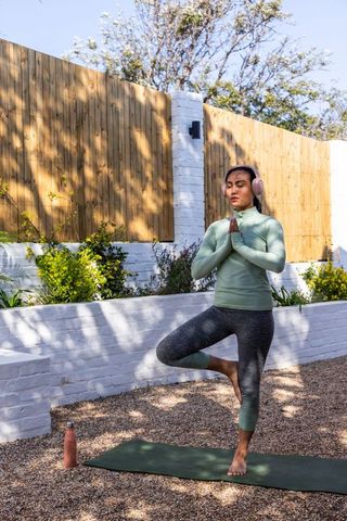 Asian woman practicing tree pose in backyard yoga with headphones and water bottle