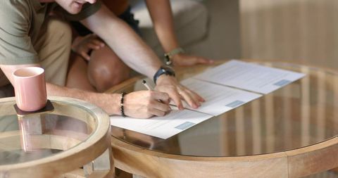 Young couple signing contract documents on glass coffee table closeup of hands pen and mug