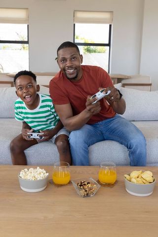 Black father and son gaming on sofa with snacks and orange juice