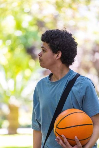 African american young man holding basketball and bag in park, casual athletic portrait