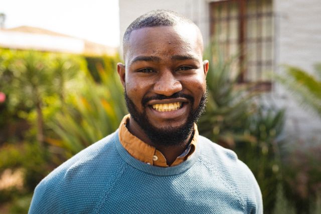 Portrait of a smiling bearded African American man standing outdoors on a sunny day. He is wearing a blue sweater over a brown shirt, exuding confidence and happiness. The background features lush greenery, suggesting a natural and relaxed environment. This image is ideal for use in lifestyle blogs, advertisements promoting positivity and confidence, or any content related to outdoor activities and casual fashion.