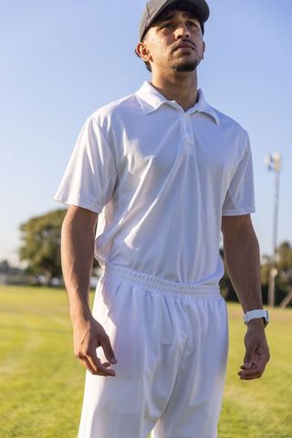 Indian male athlete standing looking up on sunlit grass field wearing white kit, cap