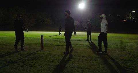 Night cricket players gathering around wicket under floodlights casting long shadows