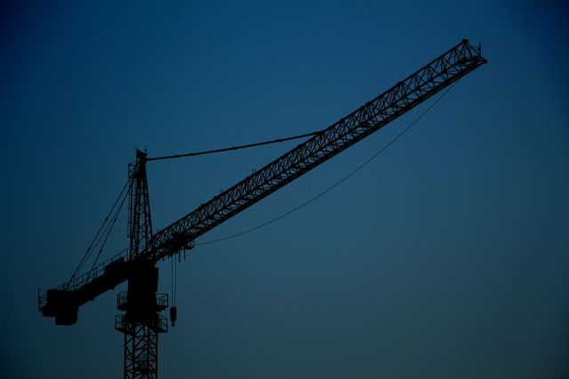 Silhouette of a construction crane stands tall against the twilight sky, showcasing the machinery and equipment used in urban development. This can be used in content related to construction, industrial work, urban development, and architecture. Perfect for illustrating the themes of engineering, building progress, and the functionality of heavy machinery during different times of the day.