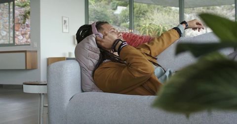 Young man relaxing on modern sofa listening to music with pink headphones and smartwatch