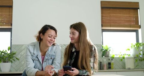 Diverse female friends sharing smartphone and laughing in bright modern kitchen