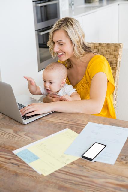 A mother is using a laptop while holding her baby in a kitchen. This image can be used to illustrate concepts of multitasking, work-life balance, remote work, and modern parenthood. It is ideal for articles or advertisements related to parenting tips, home office setups, family life, and technology use in daily life.