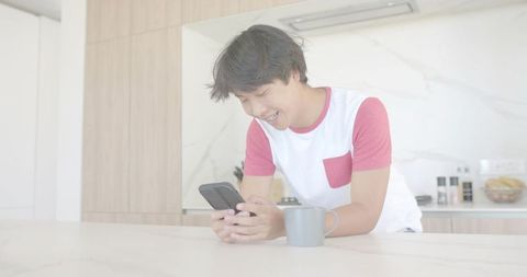 Young Asian Man Leaning on Kitchen Counter Smiling and Checking Phone with Mug