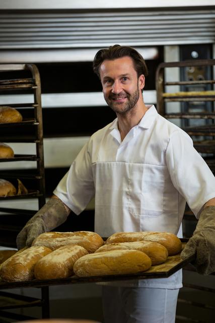 Smiling baker holding a tray of freshly baked bread in a professional kitchen. Ideal for use in articles or advertisements related to baking, culinary arts, small businesses, and food preparation. Can also be used for promoting bakery products, cooking classes, or artisan bread.