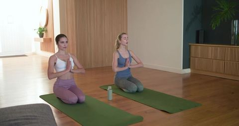 Two women practicing yoga and mindful breathing on mats in sunlit living room