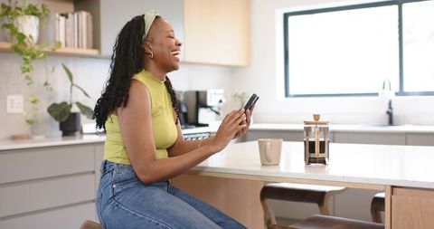 African american woman laughing while using smartphone at bright modern kitchen island