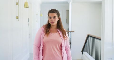 African american woman standing in sunlit modern hallway wearing pink jacket