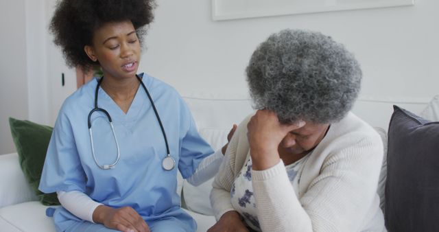 Nurse in blue scrubs with stethoscope comforting elderly woman sitting on couch, displaying support and care. Woman appears distressed, emphasizing themes of understanding and compassion in healthcare. Suitable for use in healthcare websites, senior care promotions, medical blogs, and presentations on elderly support.