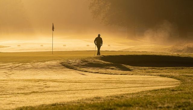 Golfer silhouette walking to flag holding putter on dew-covered green at golden sunrise
