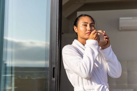 Asian woman sipping morning coffee on balcony in white bathrobe, sunlit calm and cozy