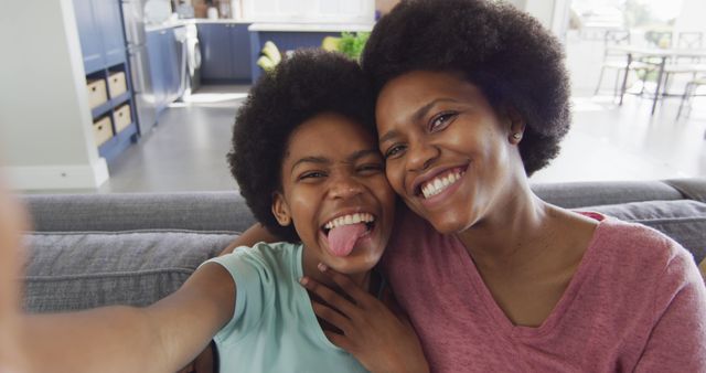 Happy african american mother and daughter sitting on sofa using smartphone, taking selfie. domestic life and quality family time together at home.