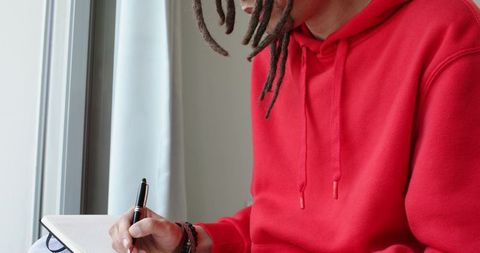 Man in red hoodie writing in notebook by sunlit window, dreadlocks, creative focus