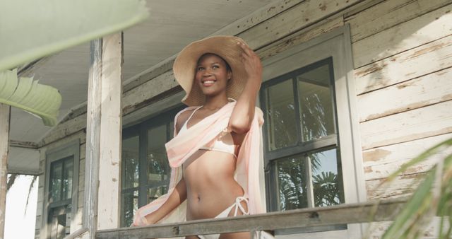 Woman wearing a bikini and a large straw hat, smiling while standing on a rustic porch. Great for summer fashion, vacation lifestyle, and outdoor leisure themes.