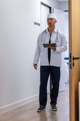 Asian physician walking clinical corridor holding tablet and stethoscope near reporting room