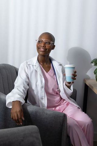 Relaxed smiling healthcare professional holding coffee cup wearing pink scrubs and white coat
